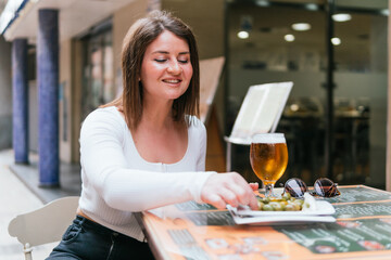 Smiling young lady sipping a beer and snacking on olives while reading a menu outdoors