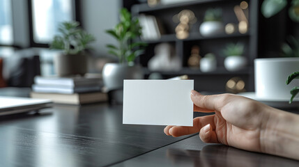 Obraz premium Hand Holding Blank Business Card in Modern Office Environment with Green Plants and Shelves