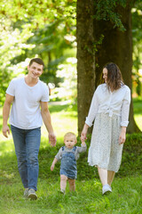 Fototapeta premium Mom and dad hold their child hands while walking along grass in park on summer day. Happy family lifestyle concept.Happy family walking in nature,feeling of fun while spending time together.Vertical