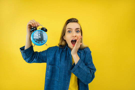 pretty girl hold alarm clock with open mouth and funny look isolated yellow color background