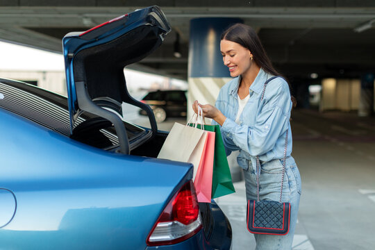 Happy woman packing her paper bags into car trunk while standing in underground parking lot of shopping mall