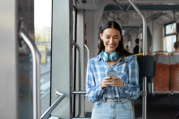 Happy European woman with headphones using cellphone, training in modern tram, enjoying comfortable...