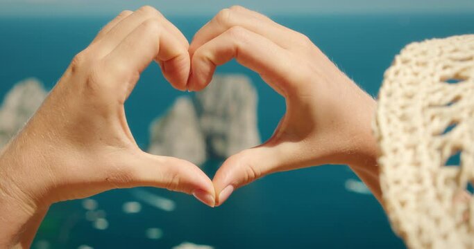 Woman shapes heart with hands over Faraglioni rocks. Romantic gesture with sea and cliffs below on Capri island, Italy.