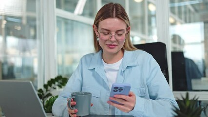 Happy caucasian businesswoman enjoying drink in blue mug while using wireless smartphone for browsing social medias indoors. Smiling young female staying in touch during coffee break in cabinet.