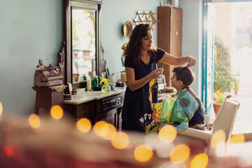 A boy is sitting in a chair, smiling, as a hairdresser trims his hair inside a brightly lit salon.