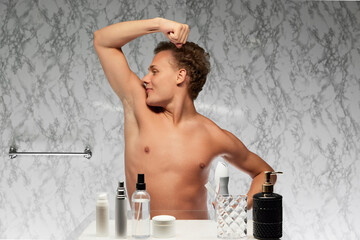 young man against marbled wall in bathroom engaged in his grooming routine, raising arm indicating...