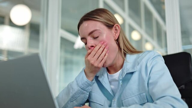 Drowsy caucasian woman yawning while taking break at private workstation surrounded by functional tools. Fatigued young professional finding respite in brightly illuminated corner of modern office.