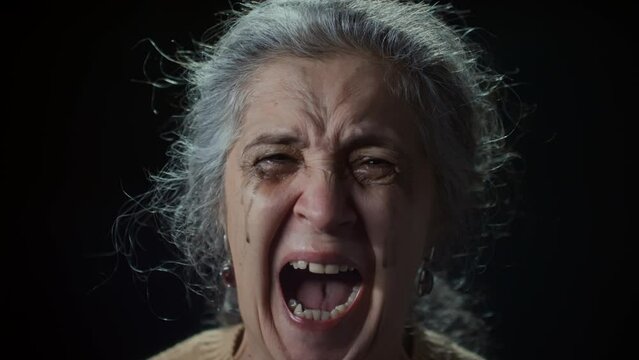 Closeup studio portrait of furious senior Caucasian lady with dripping mascara looking at camera and yelling continuously against black background, effect of shaking camera