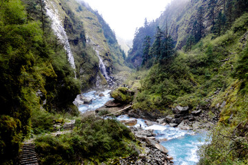 Waterfall among the himalaya mountains with green forest and a river full of rocks.