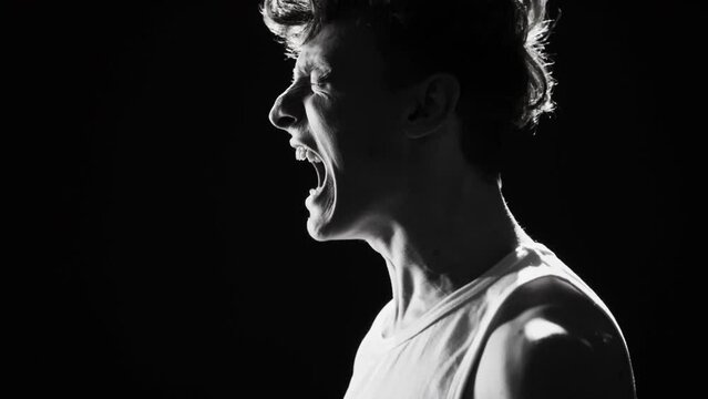 Monochrome studio portrait in profile of furious young man in undershirt shouting at top of his voice against black background