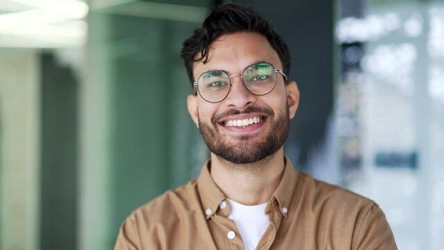 Portrait of a happy bearded manager standing in a business office. Positive smiling man worker posing looking at camera indoor. Head shot of confident handsome employee in glasses and shirt. Close up