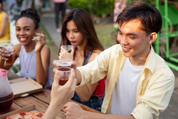 Young college-age friends making a toast and enjoying pizza during a picnic outside in the yard. A joyful gathering with smiles and shared moments.