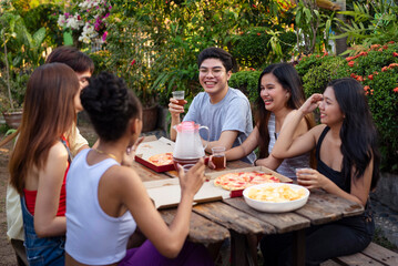 Six young friends hanging out, laughing and having a nice picnic outdoors by the backyard enjoying pizza and drinks