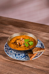 bowl of pumpkin orange soup with croutons, cilantro leaves and sesame seeds toppings, over ceramic blue flower patterned plate on rustic wooden table 