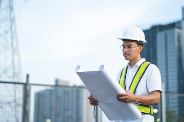 A man wearing a hard hat and safety vest is holding a blueprint. He is standing in front of a tall...