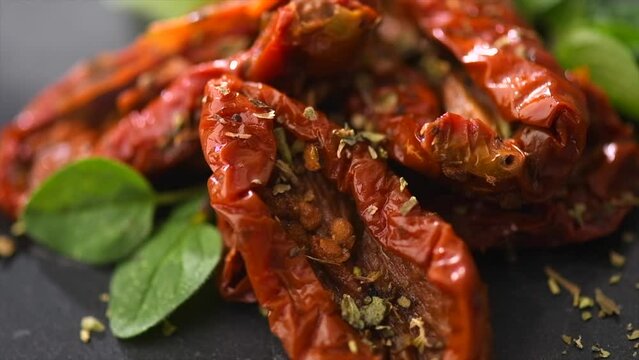 Sun dried tomatoes with aromatic herbs, oregano, marjoram leaf. Rotating on black stone background, close up. Italian cuisine, healthy vegan food, Mediterranean diet. Dry tomato. 