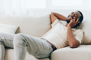 Obraz premium Happy African American man sitting on a black sofa, holding a smartphone and smiling He is engrossed in a video call, enjoying the modern technology and comfort of his home The background shows a cozy
