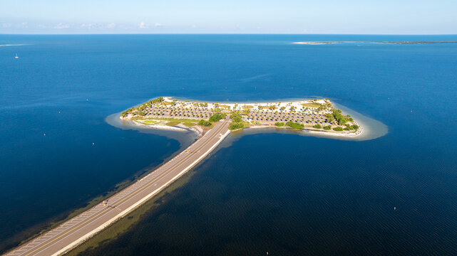 Aerial view of Fred Howard Park in Tarpon Springs, Florida, featuring parking lots, palm trees, sandy beaches, and surrounding blue waters under a clear sky.