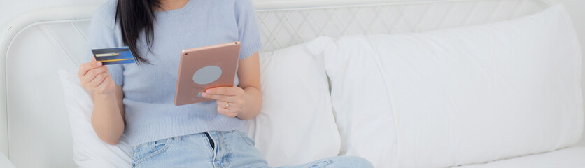 Young asian woman sitting on bed using digital tablet shopping online with credit card in the...