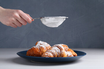 Woman with sieve sprinkling powdered sugar onto croissants at white wooden table, closeup