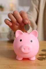 Woman putting coin into pink piggy bank at wooden table, closeup