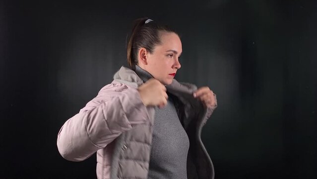A beautiful young woman puts on a coat, wearing a gray sweater with an air of casual look, her piercing gaze exuding an edgy yet confident attitude against the minimal black backdrop