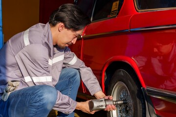 Asian male mechanic kneeling and using power tool on car wheel in auto repair shop. Wearing grey...