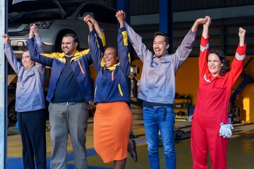 male and female mechanics holding hands, smiling, celebrating success in auto repair shop. Wearing different uniforms, teamwork, and positive work atmosphere in professional services.