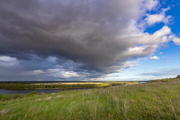 A serene meadow overlooks a winding river under a dramatic sky filled with storm clouds and patches of blue, capturing the beauty of nature's ever-changing weather patterns.