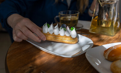 A hand delicately picks up a cream-topped eclair with a blueberry on a white plate, set on a wooden...