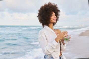 A woman with curly hair is standing on a beach, holding a drink in a plastic cup