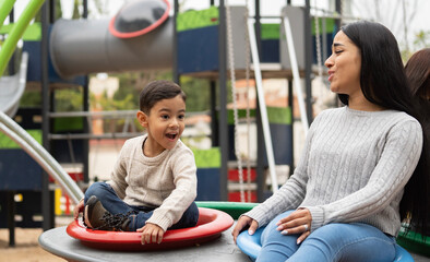 Latin mother and her son are enjoying together on red and blue wheel in a playground as a weekend activity