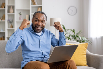 A man in a blue shirt celebrates success on his laptop at home, showing excitement and raising his...