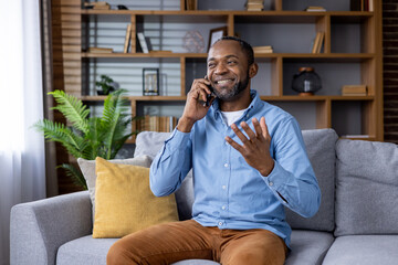 A cheerful man is engaging in a phone call while relaxing on a couch in a modern living room, exuding a happy and friendly demeanor in a comfortable and cozy home environment