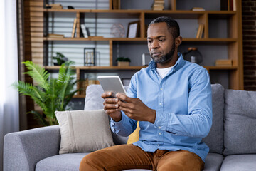 A focused man is seen sitting on a couch and working on a tablet in a modern living room. He...
