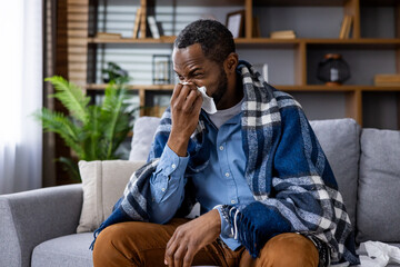 A man is sneezing in the living room, wrapped in a plaid blanket, portraying a scene of home rest...