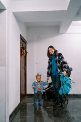 Smiling mother holding the hand of a little girl standing with a stroller in front of the door of...