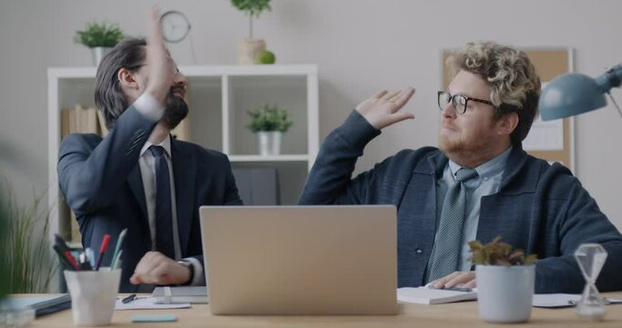 Loop of two businessmen colleagues doing high-five hand gesture enjoying cooperation sitting at desk in office. Teamwork and business concept.