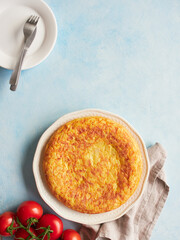 Homemade potato omelette, typical Spanish food called tortilla de patatas, with fresh red tomatoes on blue background.