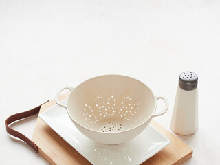 Empty colander on white plate and wooden cutting board with leather handle, next to the salt shaker on the table with copy space.
