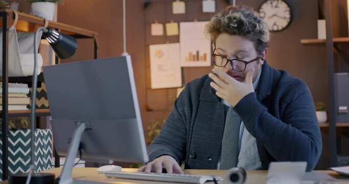 Loop of tired office worker yawning feeling sleepy while working with computer at desk in dark workplace in evening. People and job concept.