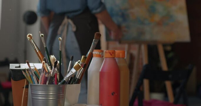 Close-up slow motion of female hand taking paintbrush then female artist walking to picture on easel busy with painting in studio. Fine arts and people concept.