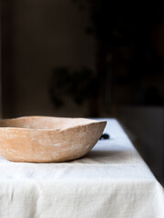 An empty ceramic plate on a white linen tablecloth against a dark blurred wall, front view. A place to record. The plate is brown, natural color. Natural clay.