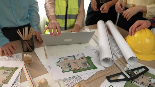 Architect and safety engineer discussion about building blueprint displaying on laptop on table with architectural document and wooden block scatter around at modern office. Closeup. Delineation.