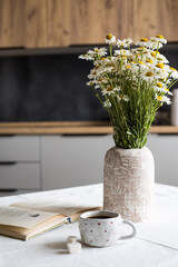 A cup of coffee, two pieces of sugar, a vase with wildflowers and an open book on the table against the background of the kitchen interior.