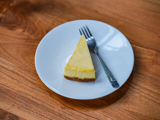 A slice of cheese cake with white plate and fork, in top of wooden table
