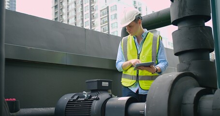A Engineer man looking inspecting maintenance insulated pipelines valve pump control on the roof at an industrial site. He is wearing a hard hat and safety vest