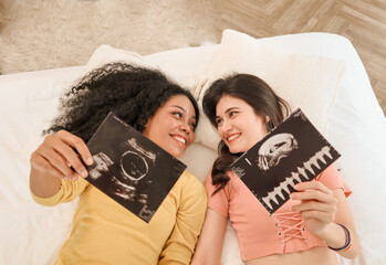 A multiracial lesbian couple lying and showing ultrasound images, two lesbians feeling excited about having their first baby by IVF (In vitro fertilization) process. Pregnancy and motherhood concept