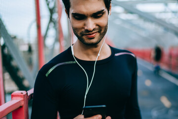 Cheerful fit man listening favourite music songs in telephone player via earphones connected to phone for workout in urban setting.Smiling jogger choosing audio application on cellular for training