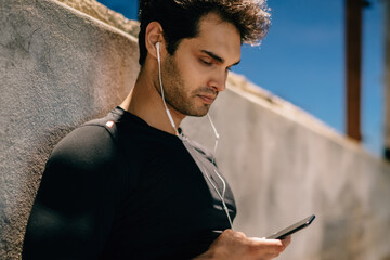 Side view of athlete dressed in active wear for training standing in urban setting with phone in...